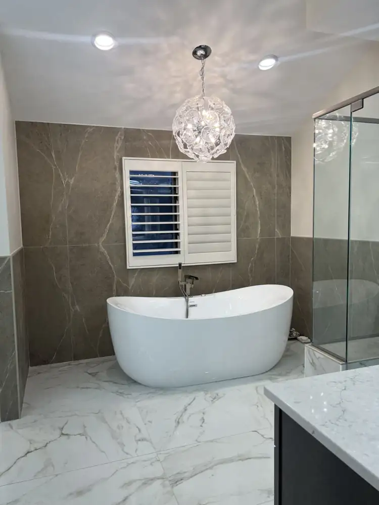 Freestanding soaking tub under a glass chandelier with large-format porcelain wall and plantation shutters