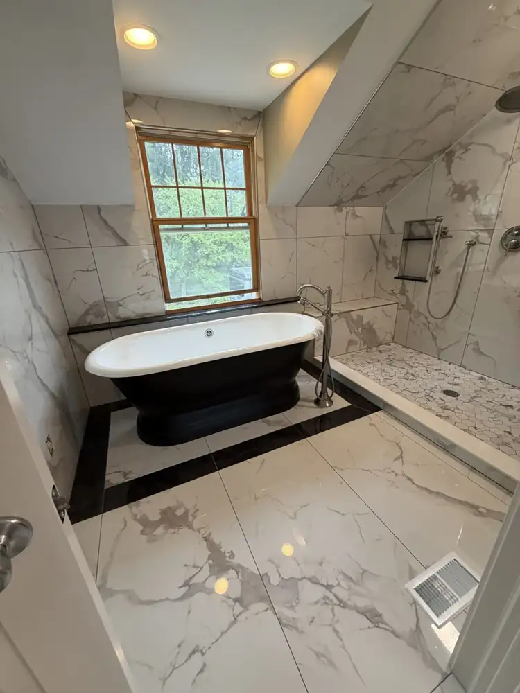 Black pedestal tub on a black-bordered marble-look tile inset under a dormer window, walk-in shower with bench, Chagrin Falls, Ohio