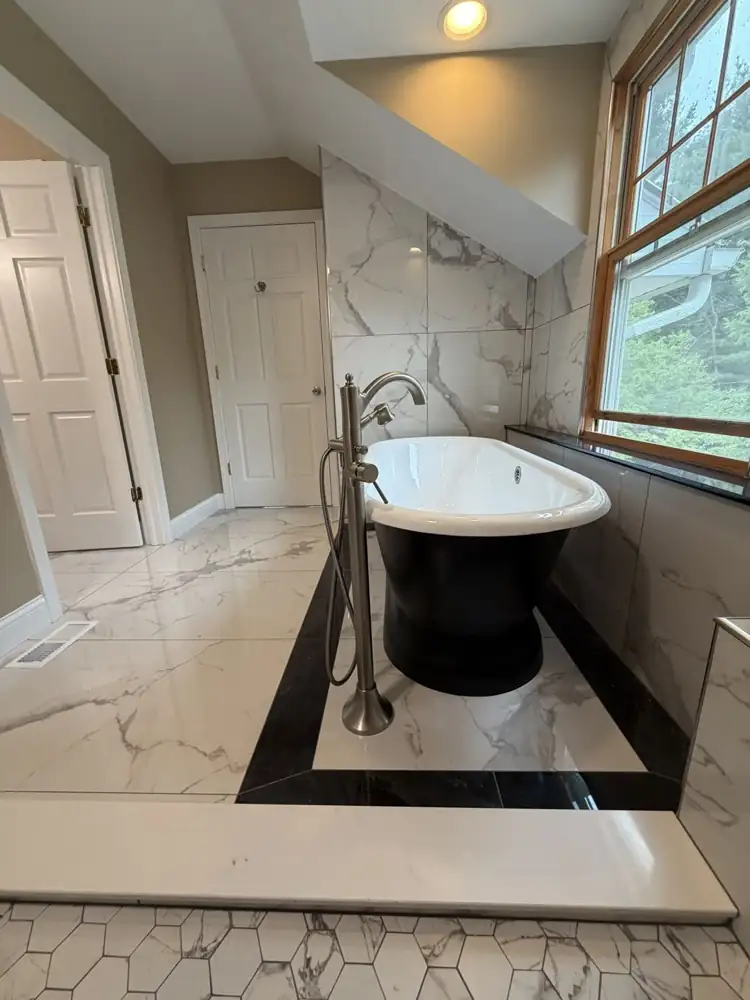 Another view: Black pedestal tub on a black-bordered marble-look tile inset under a dormer window, walk-in shower with bench, Chagrin Falls, Ohio