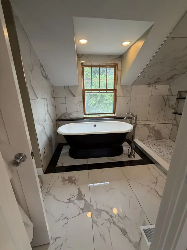 Another view: Black pedestal tub on a black-bordered marble-look tile inset under a dormer window, walk-in shower with bench, Chagrin Falls, Ohio