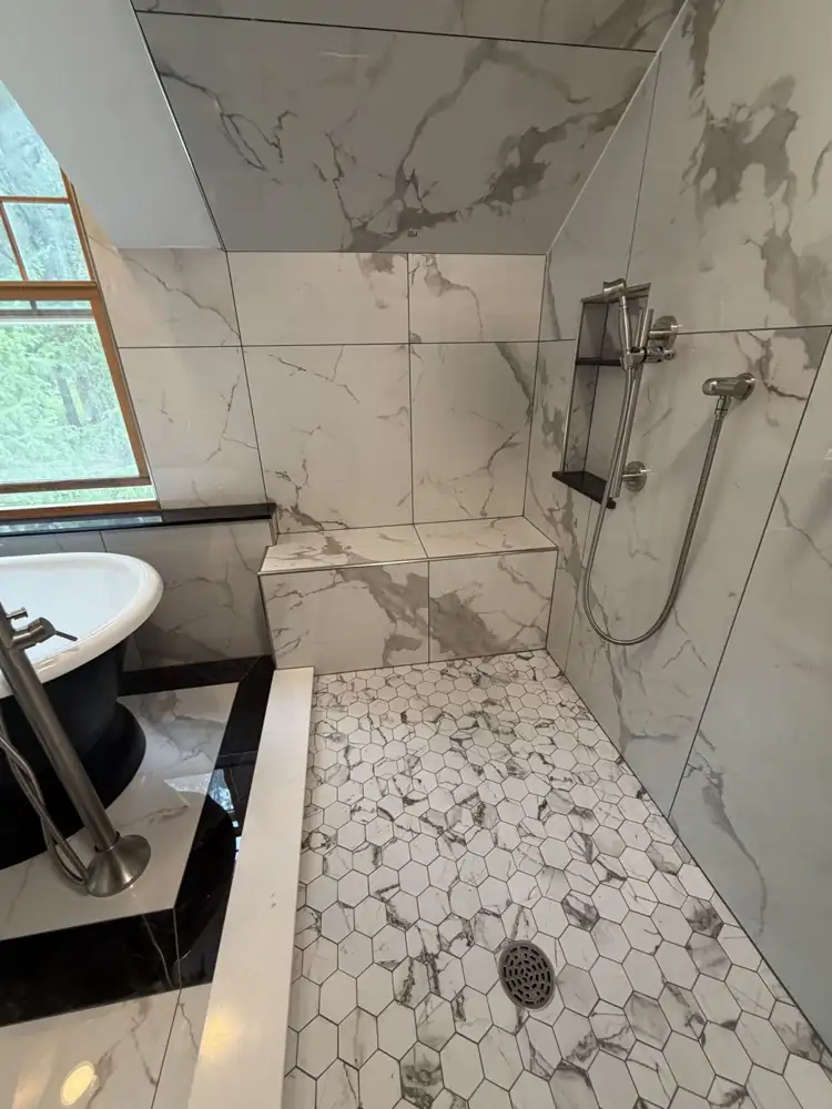 Another view: Black pedestal tub on a black-bordered marble-look tile inset under a dormer window, walk-in shower with bench, Chagrin Falls, Ohio
