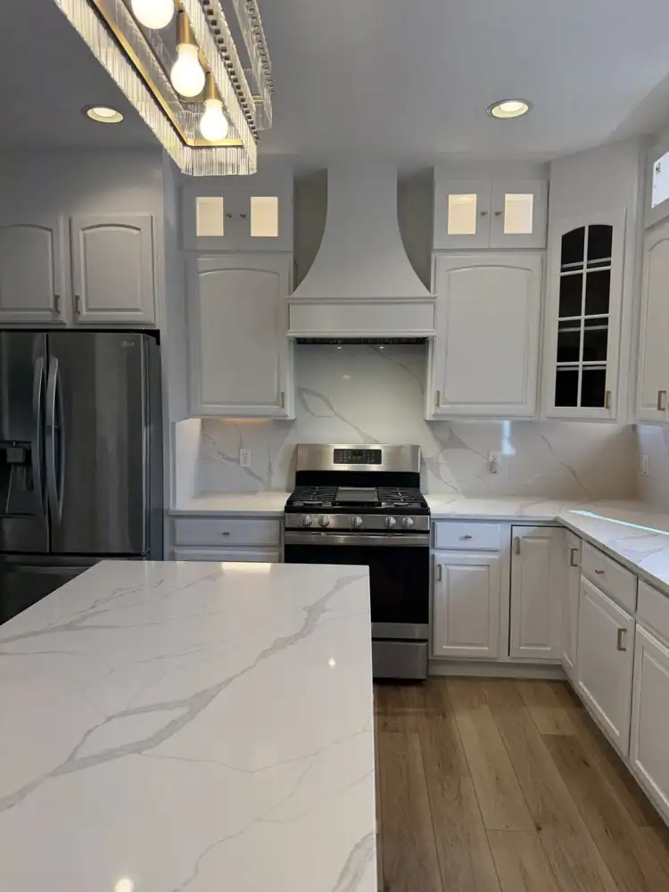 Another view: Kitchen with cabinets painted white, lit glass-front toppers, quartz waterfall island, full-height quartz backsplash, crystal chandelier and plank floor, Macedonia, Ohio