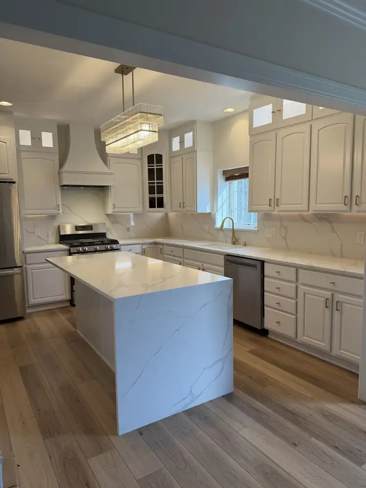 Another view: Kitchen with cabinets painted white, lit glass-front toppers, quartz waterfall island, full-height quartz backsplash, crystal chandelier and plank floor, Macedonia, Ohio