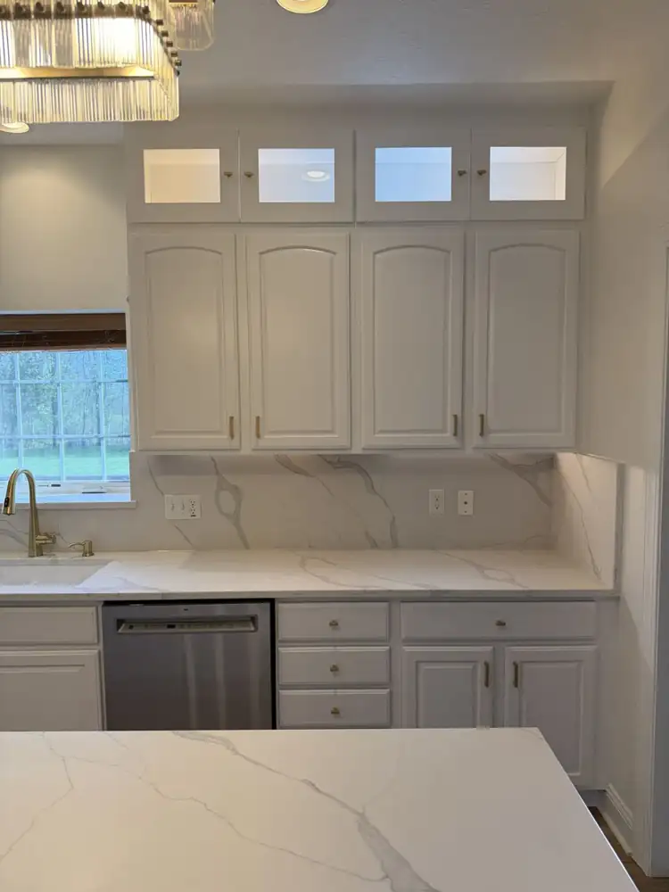 Another view: Kitchen with cabinets painted white, lit glass-front toppers, quartz waterfall island, full-height quartz backsplash, crystal chandelier and plank floor, Macedonia, Ohio
