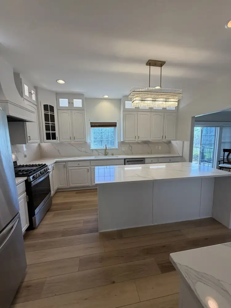 Another view: Kitchen with cabinets painted white, lit glass-front toppers, quartz waterfall island, full-height quartz backsplash, crystal chandelier and plank floor, Macedonia, Ohio