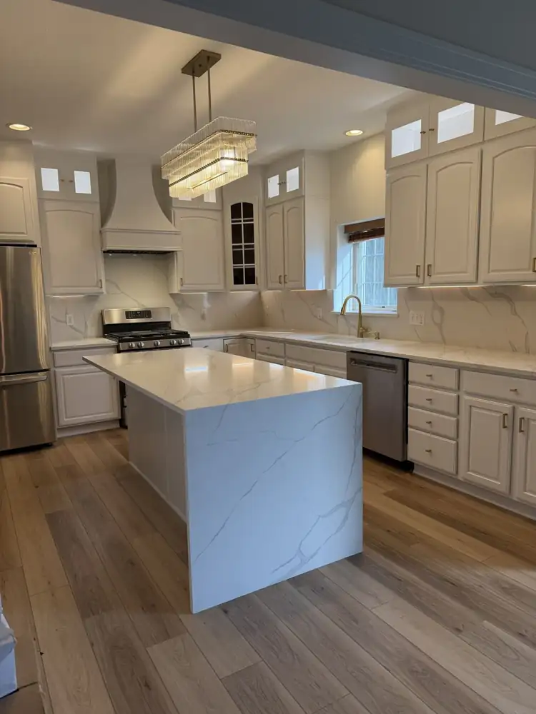 Another view: Kitchen with cabinets painted white, lit glass-front toppers, quartz waterfall island, full-height quartz backsplash, crystal chandelier and plank floor, Macedonia, Ohio