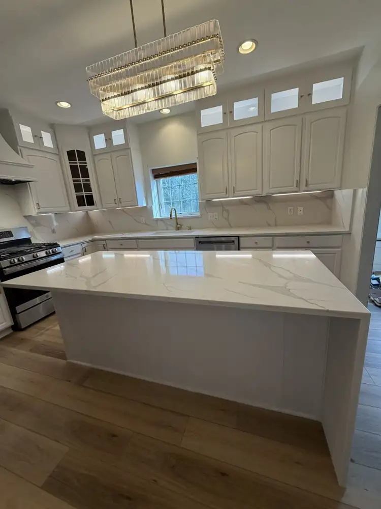 Another view: Kitchen with cabinets painted white, lit glass-front toppers, quartz waterfall island, full-height quartz backsplash, crystal chandelier and plank floor, Macedonia, Ohio