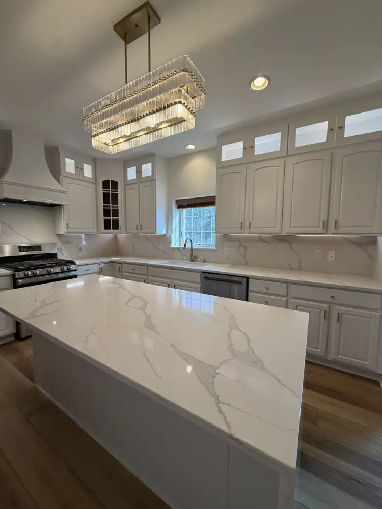 Another view: Kitchen with cabinets painted white, lit glass-front toppers, quartz waterfall island, full-height quartz backsplash, crystal chandelier and plank floor, Macedonia, Ohio