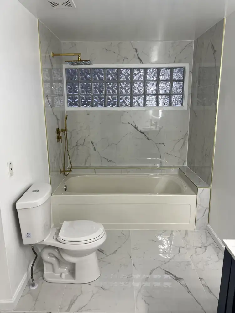 Tub with white marble-look porcelain surround, glass block window and brushed gold rain shower, Northeast Ohio