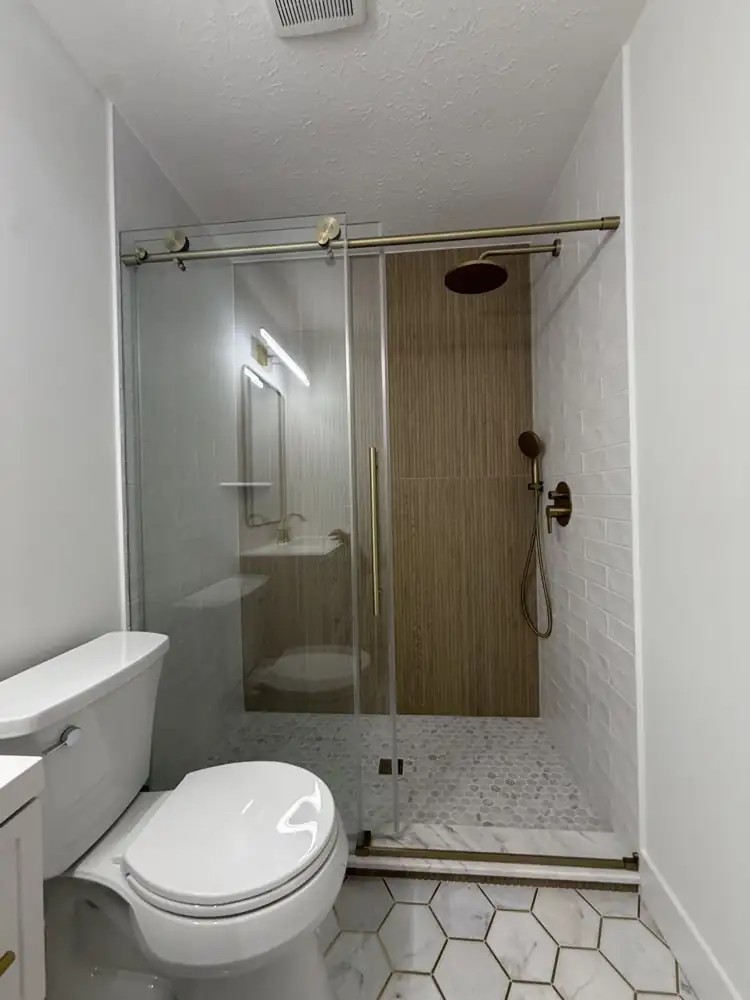 Another view: Bathroom with walk-in shower, fluted wood-look tile wall, sliding glass door, brushed gold fixtures and marble hex floor with brass inlay, Rocky River, Ohio